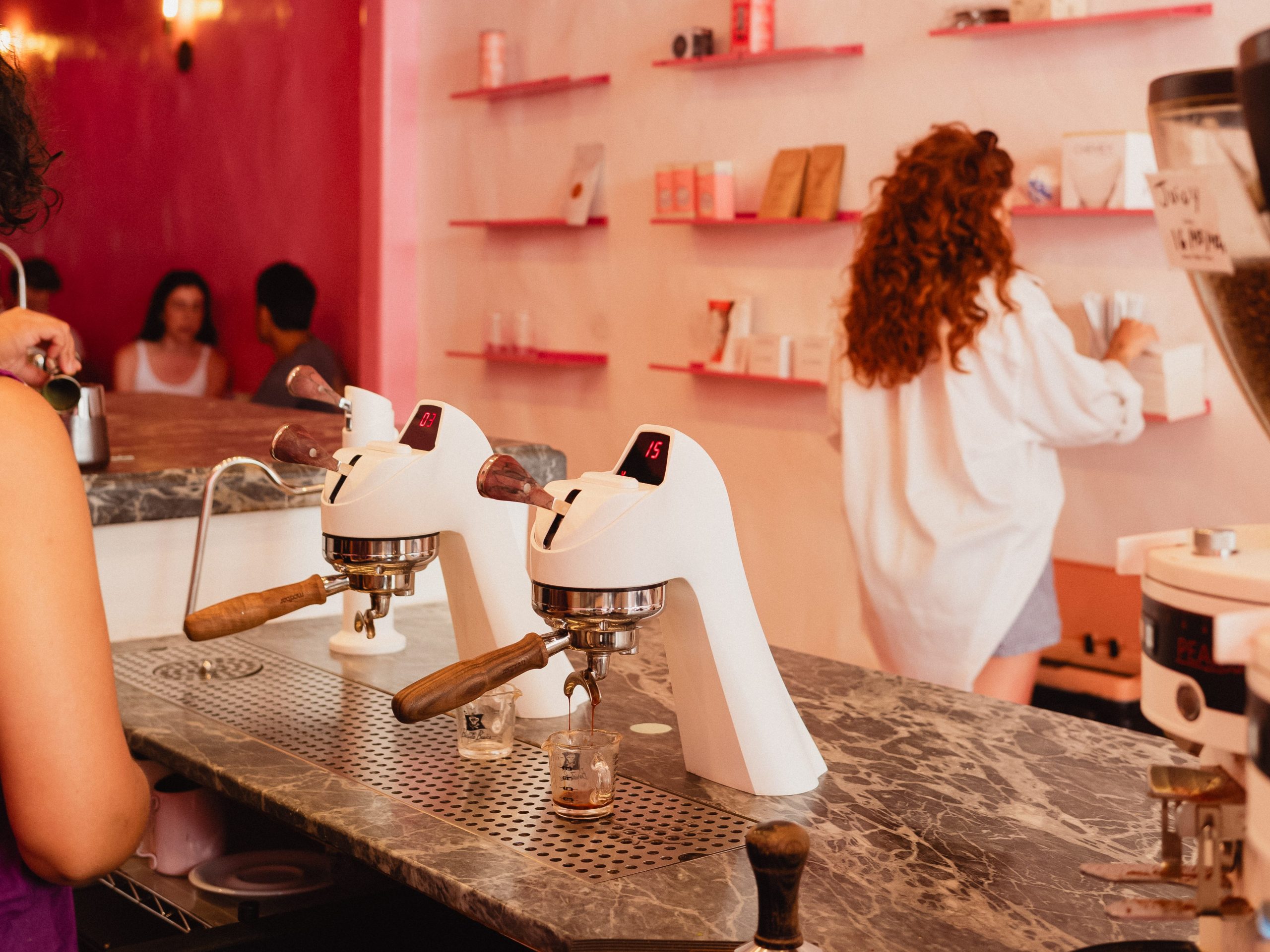 dayglow's sunset blvd cafe. from behind the counter, two white modbar taps with portafilters that have walnut handles. one is pulling a shot of espresso. a person browses coffee bags on a shelf in the background