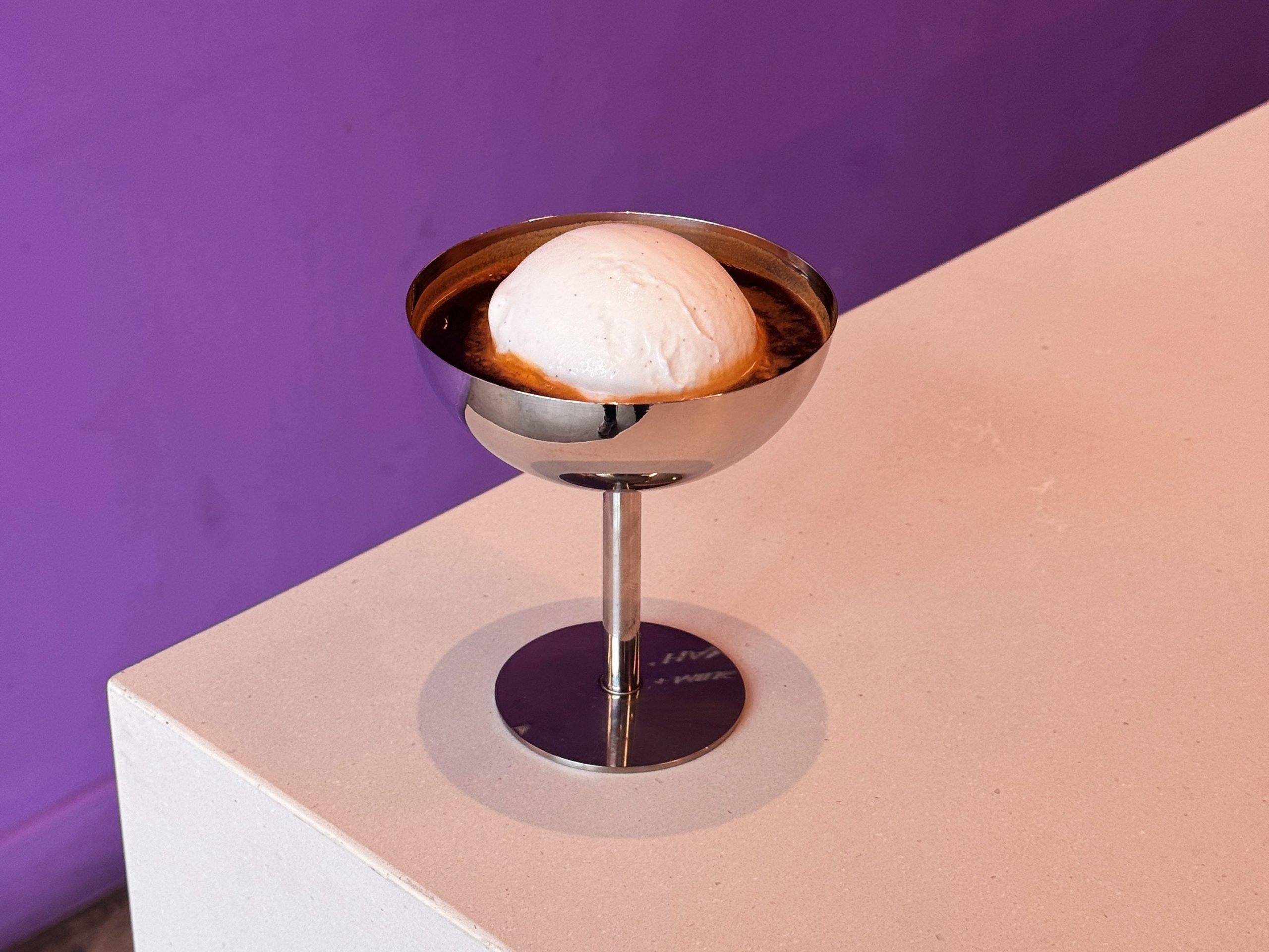 a scoop of ice cream floting in espresso in a silver goblet on a counter against a purple background