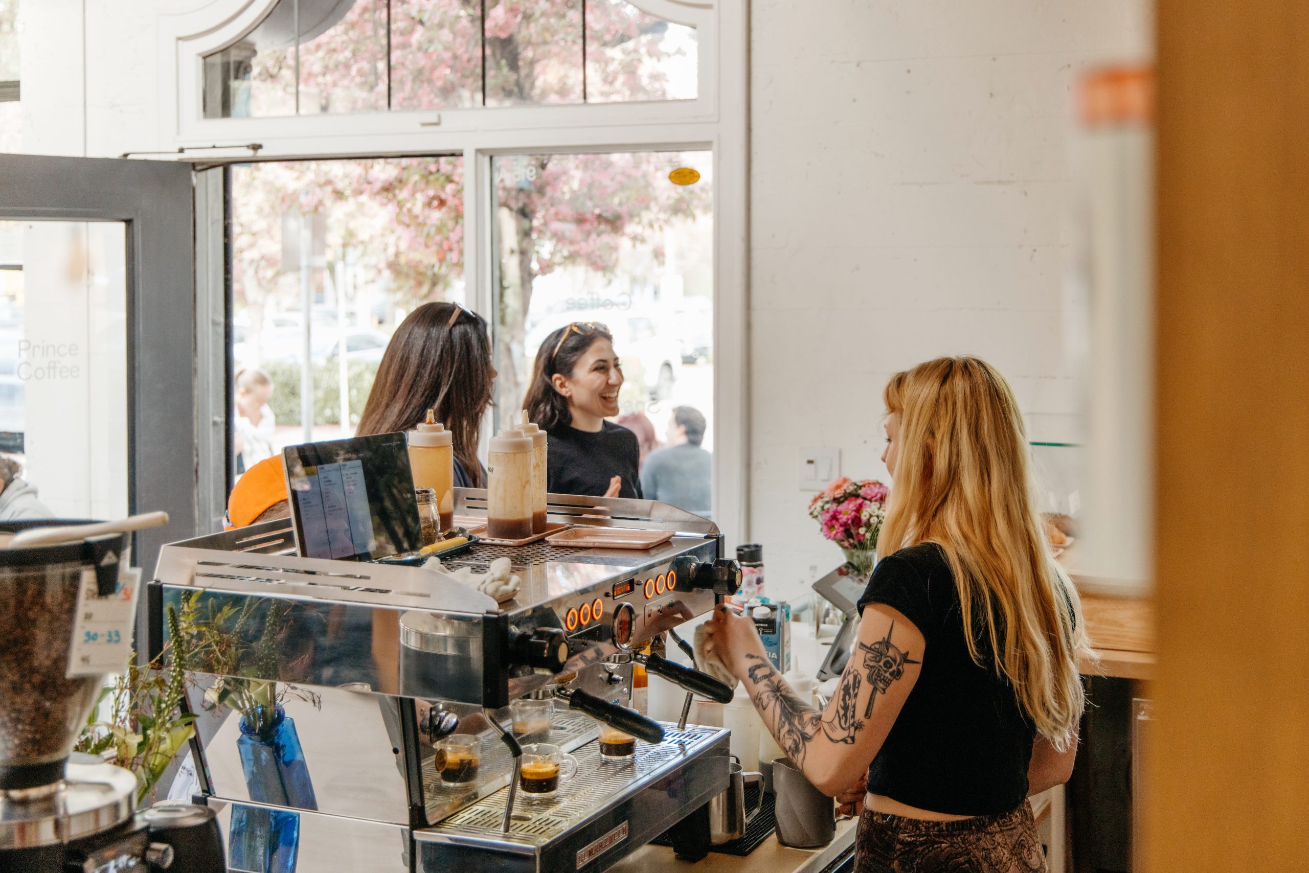 prince coffee cafe, shot of behind the bar showing the front of the espresso machine with a barista making drinks for two smiling customers